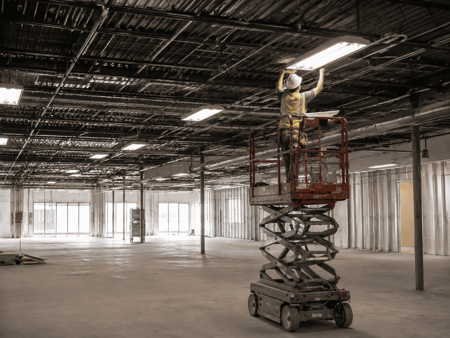 A construction worker on a scissor lift installs or repairs a ceiling fixture inside a large, unfinished commercial building.