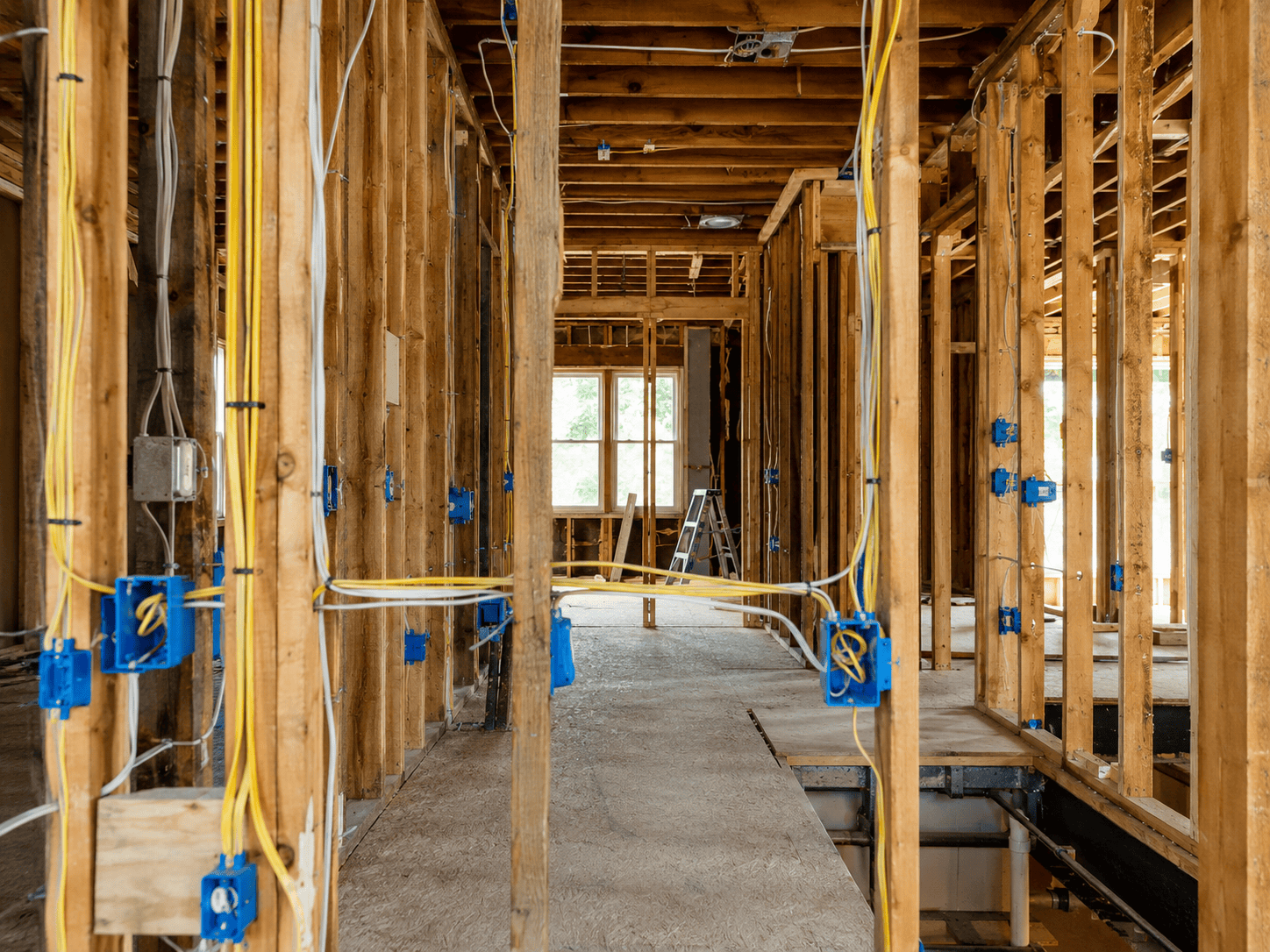 Interior of a house under construction with exposed wooden framing, electrical wiring, and blue outlet boxes; ladder near window at the far end.