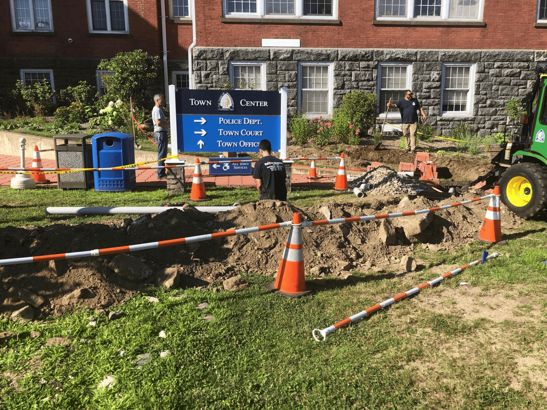 Construction area with dirt piles, orange cones, and caution tape in front of a Town Center sign and brick building; workers and a green tractor are present.